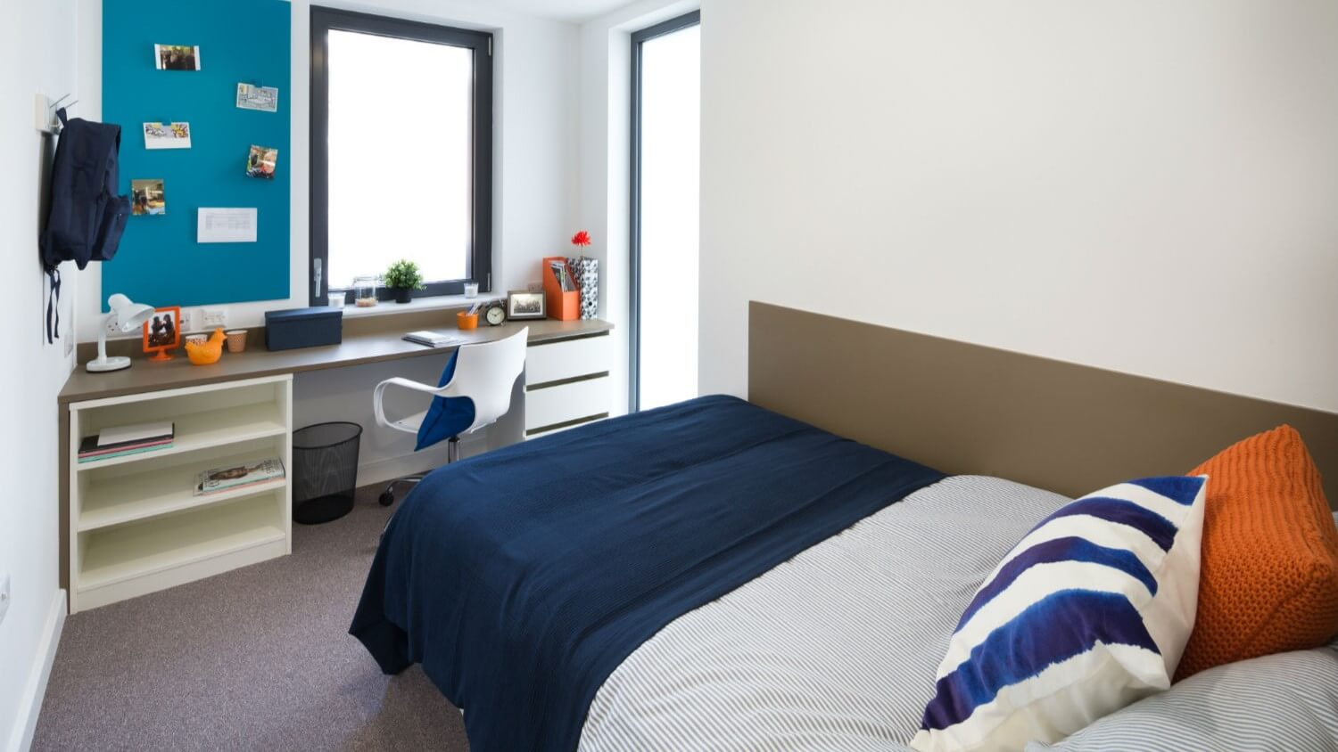 A bedroom at Grand Felda House accommodation with a bed, desk, chair, two windows and a notice board.