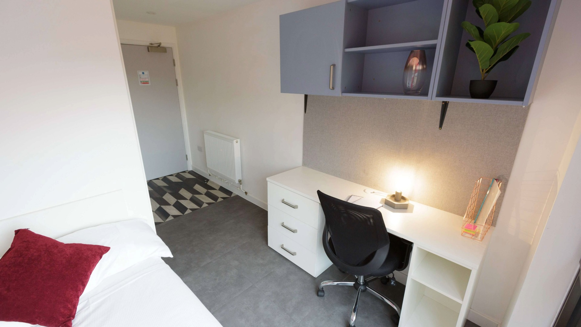A room with bed, red pillow, desk with lamp, black chair, and wall shelves.