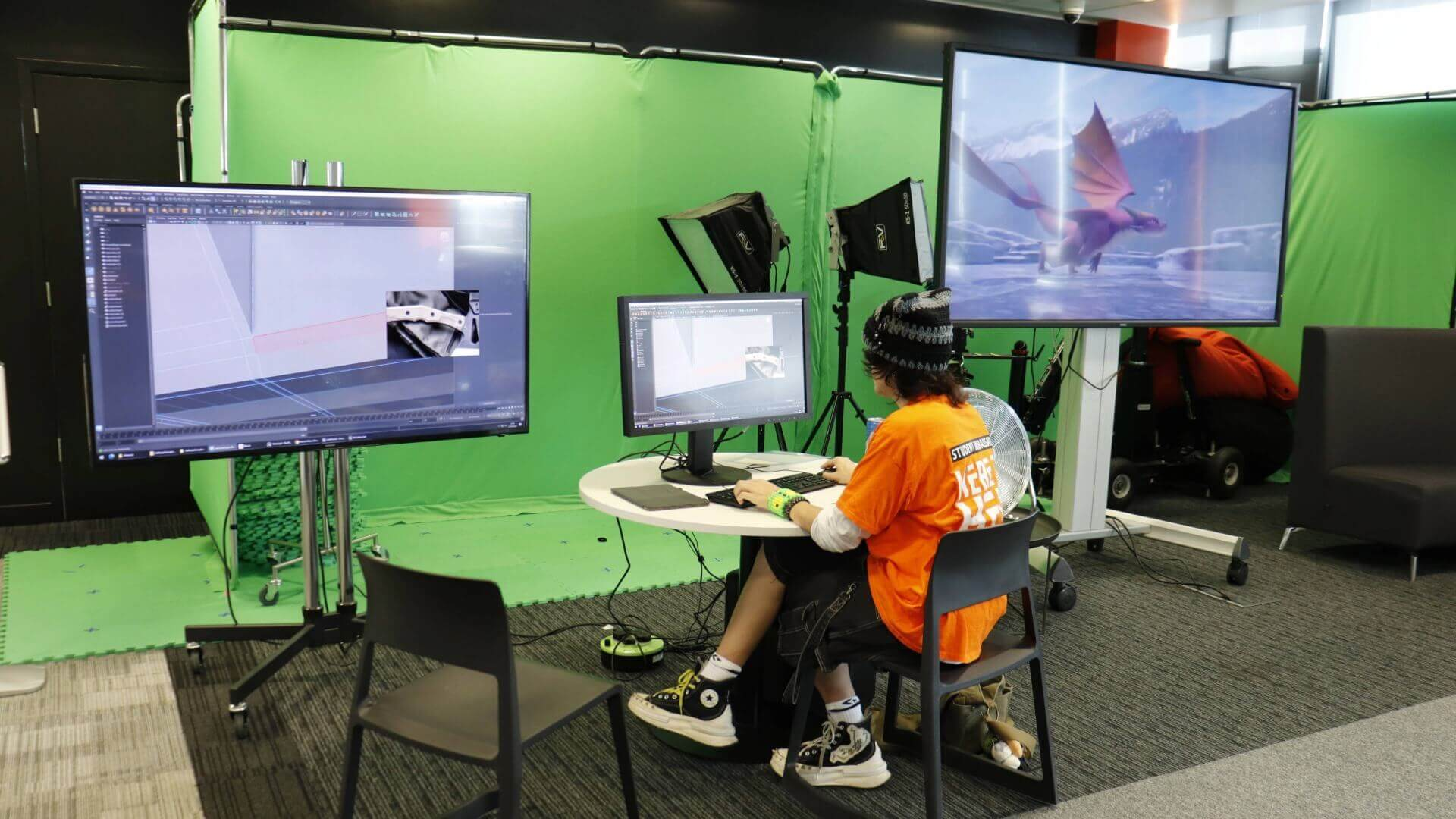A student ambassador in an orange T shirt and beanie hat works on a computer and large screen in the Escape Studios breakout space.