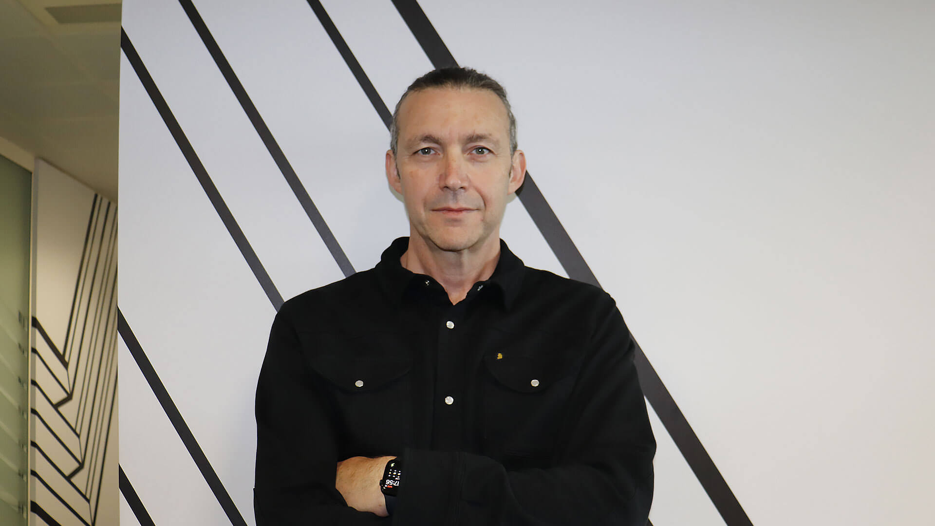 A person with a ponytail and a black shirt stands with arms crossed in front of a wall with diagonal black lines.