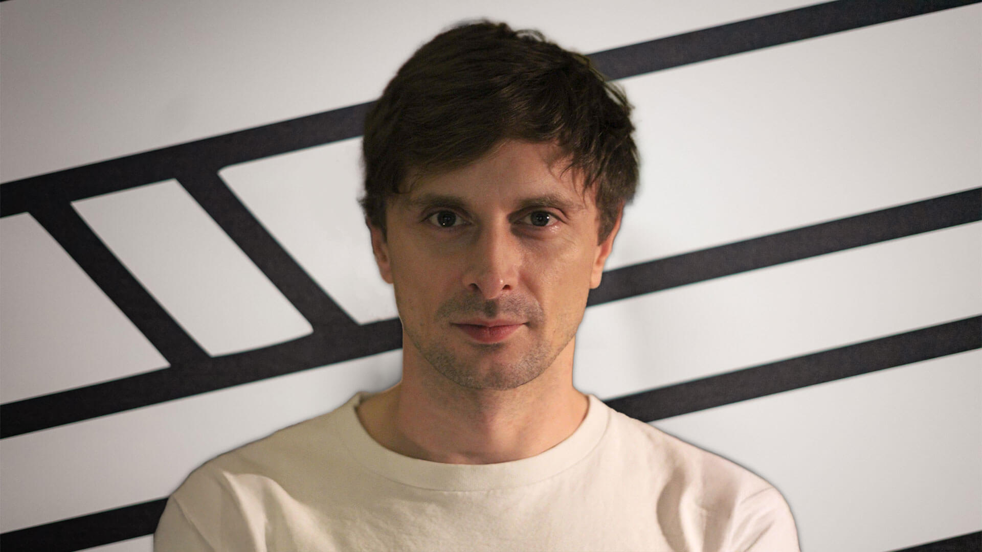 A person with short brown hair wearing a white shirt stands in front of a wall with black geometric lines.