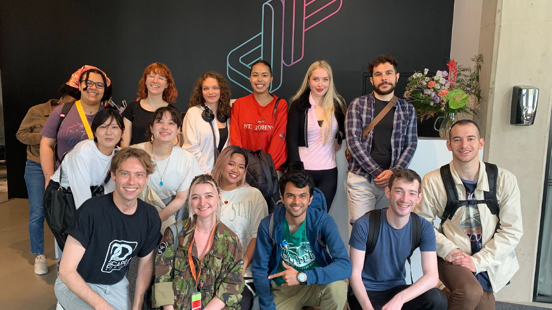 A group of recent Escape Studios graduates smiling in front of a wall with a large Jellyfish Pictures logo on it.