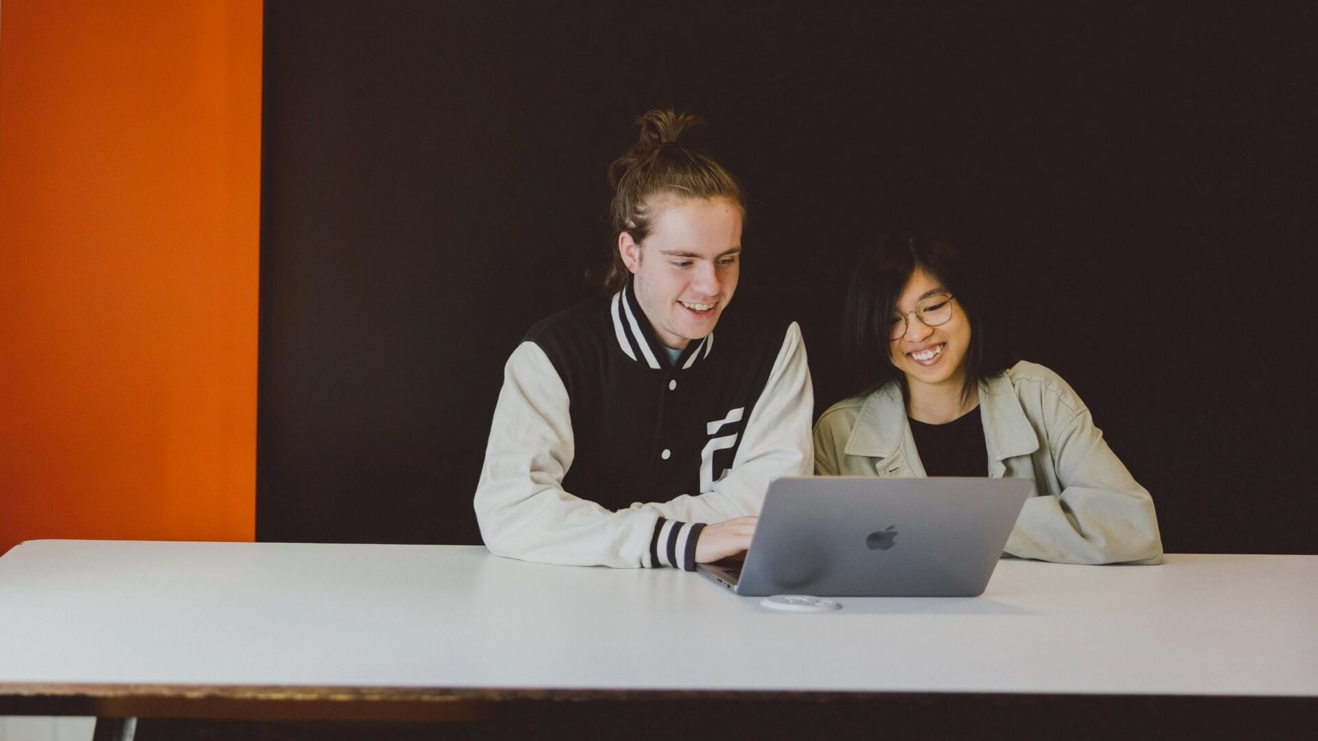 Two students sat at a table, both looking at a laptop and smiling.