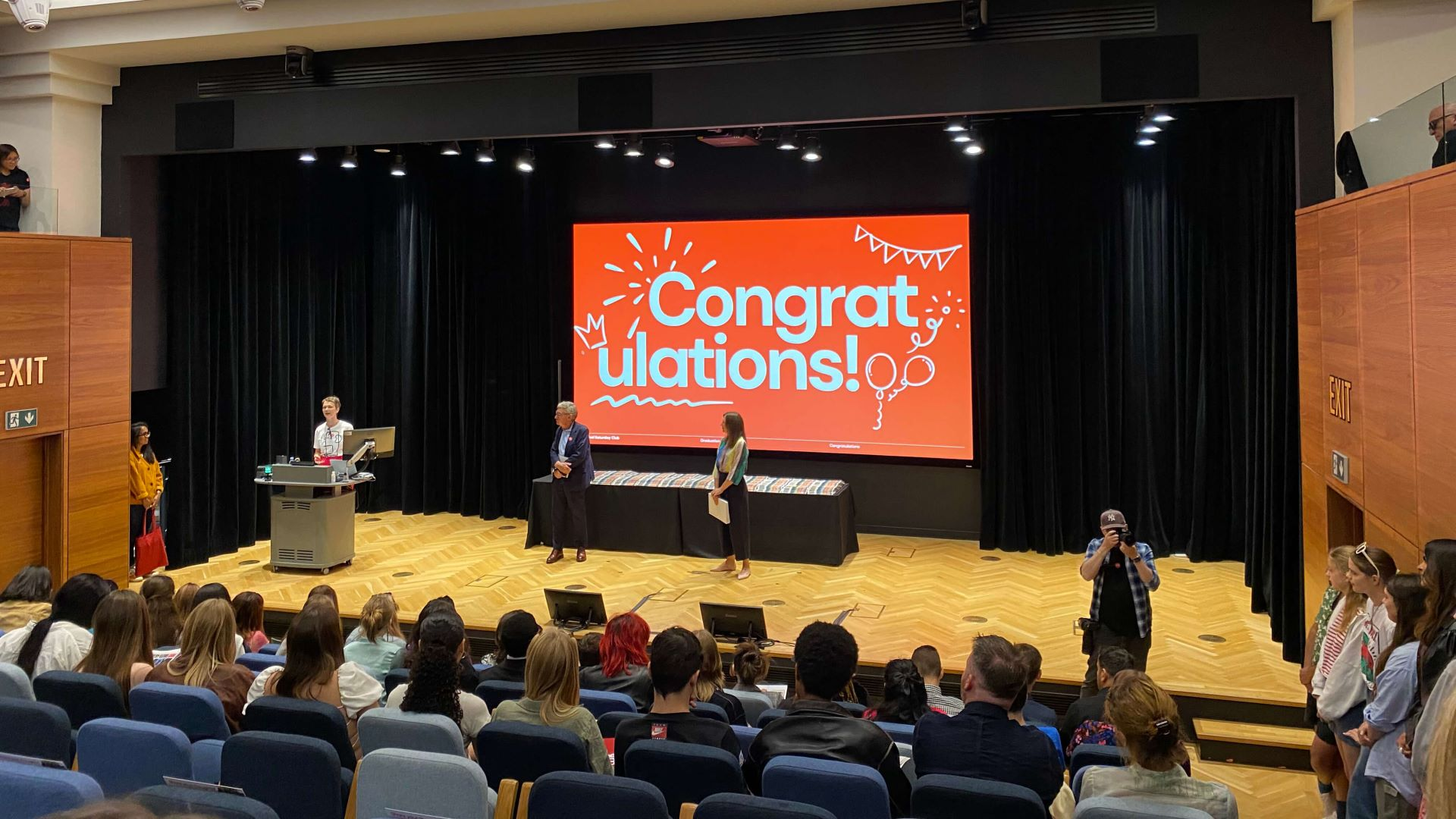The members of the Saturday club waiting to graduate in theatre seating facing a satge. On stage, there are two speakers, and a sign saying 'Congratulations!'!