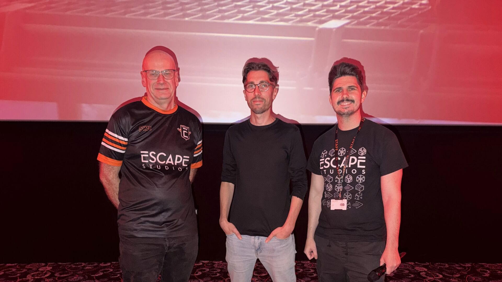 Three men standing indoors, two wearing Escape Studios T-shirts. The background is dimly lit with a red tone.
