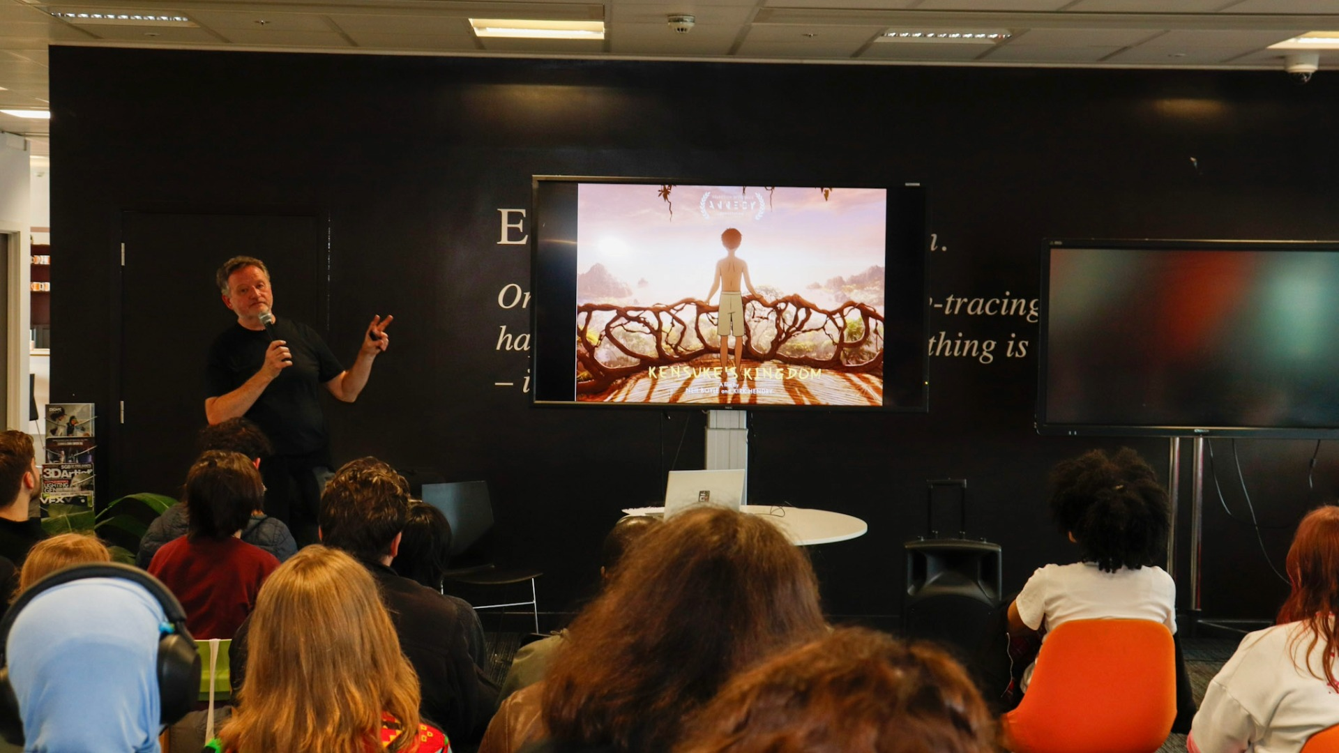 Kensuke's Kingdom director Neil Boyle holds a microphone and presents slides on a screen to a student audience.