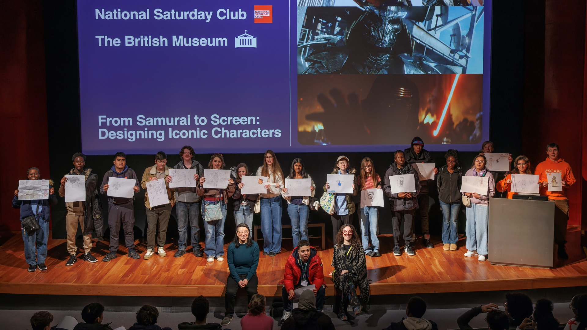 Group photo of Escape Studios Film and Screen Saturday Club members holding up drawings done during an activity on a stage.