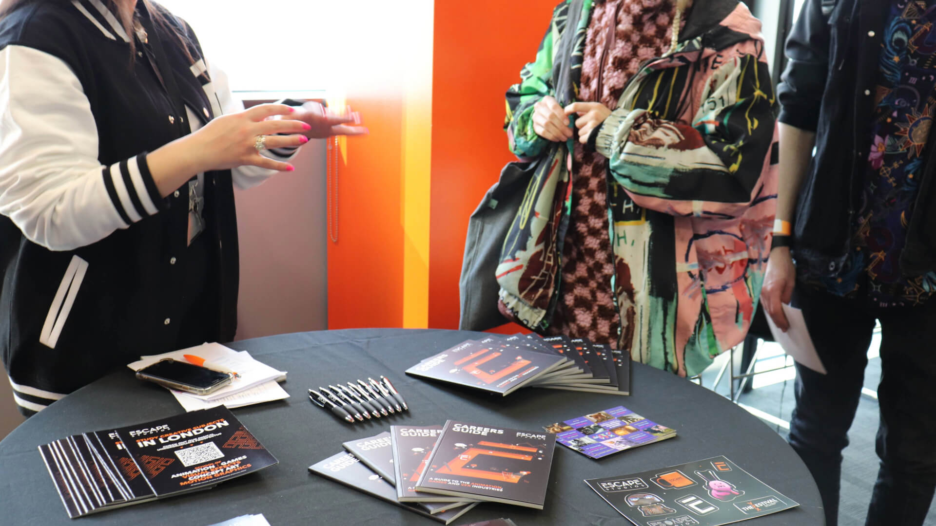 An Escape Studios staff member chats to two open day attendees over a table displaying Escape studios leaflets, pens and stickers