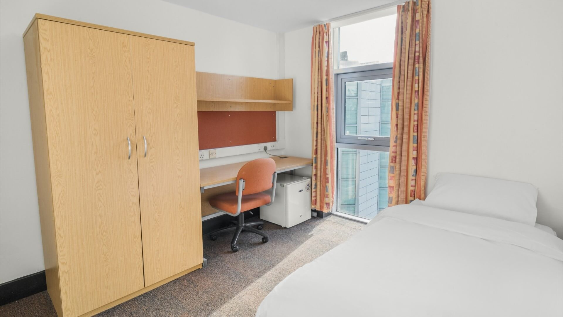 An ensuite bedroom at Davies Court with a single bed, wardrobe, desk with chair and mini fridge next to a large window with orange curtains