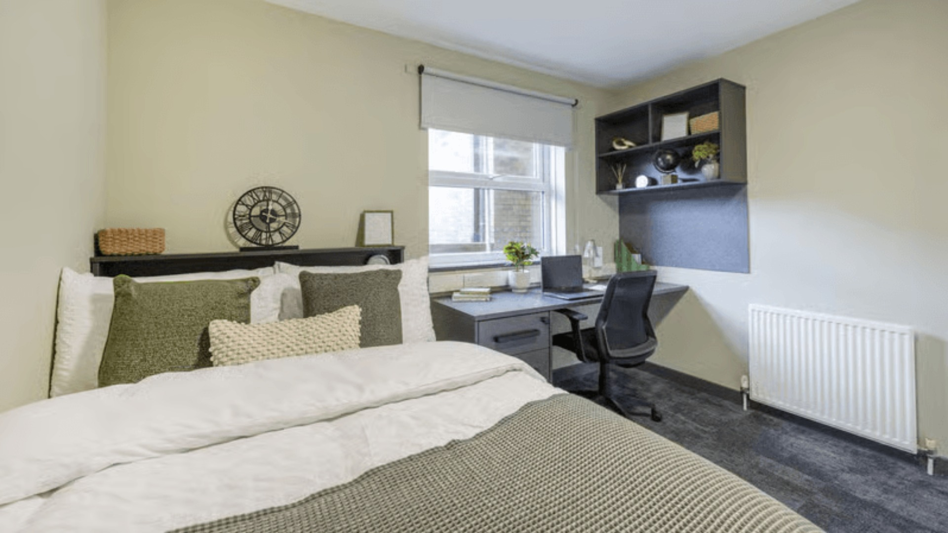 A bedroom with a bed, green pillows, a workstation with a laptop, shelves, and a window with blinds.