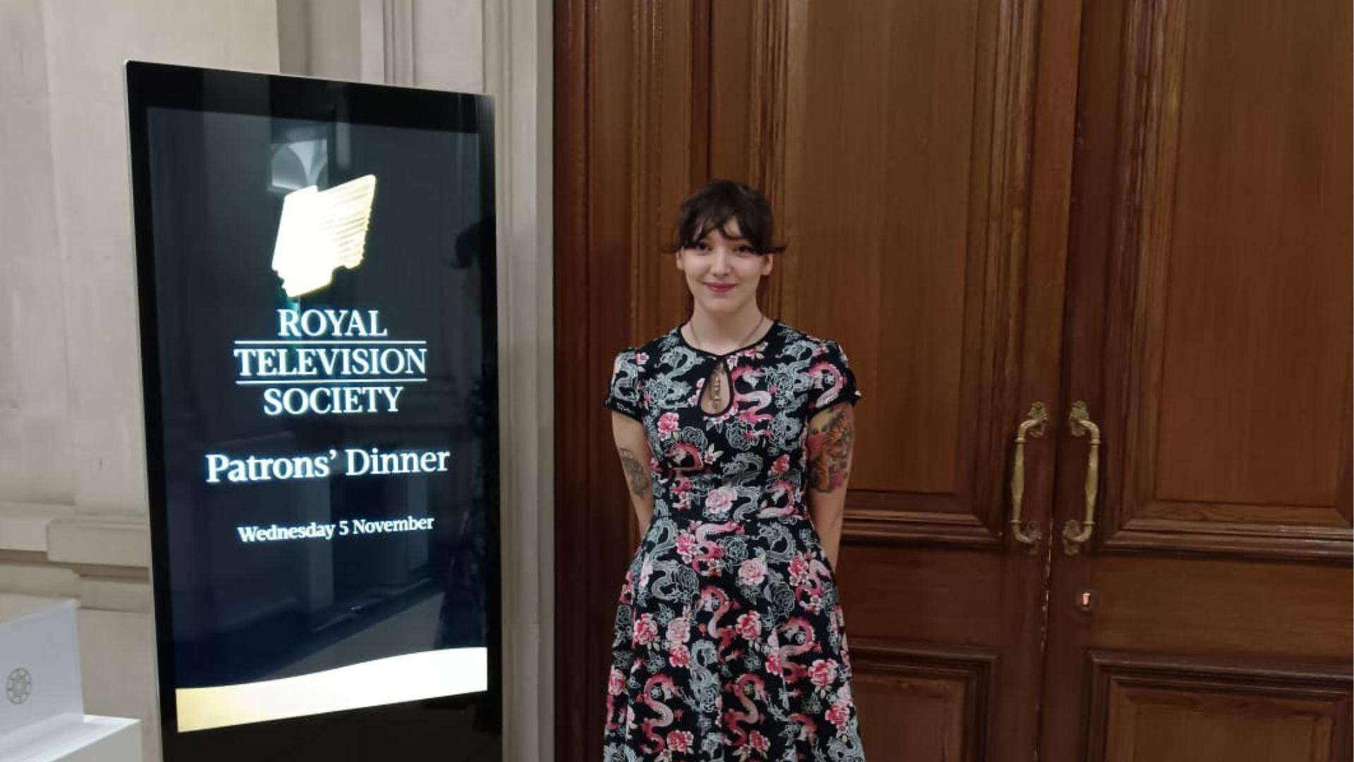 Jesse at a Royal Television Society event. She is dressed smartly and stands next to a sign reading: 'ROYAL TELEVISION SOCIETY, Patrons' Dinner, Wednesday 5th November'.