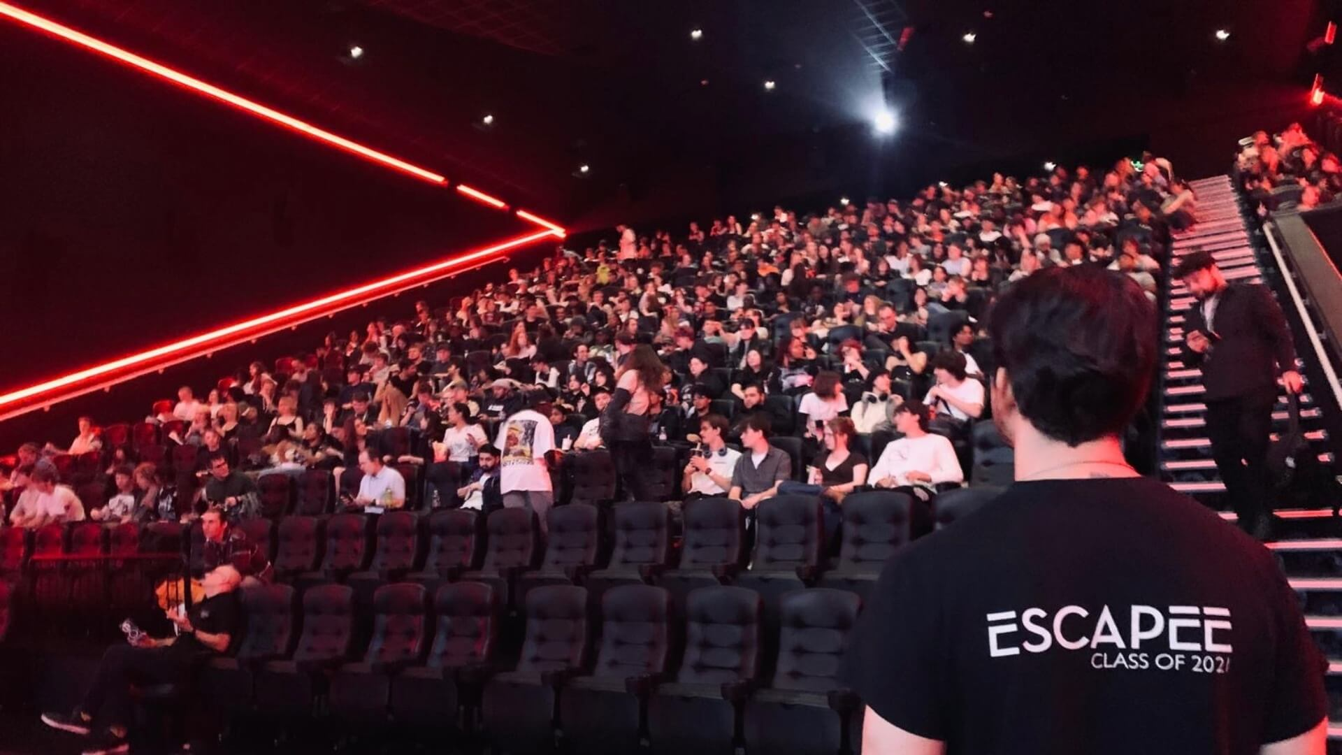 students fill the seats in a large cinema screen. A staff member wearing a branded 'Escapee' T shirt stands facing them.