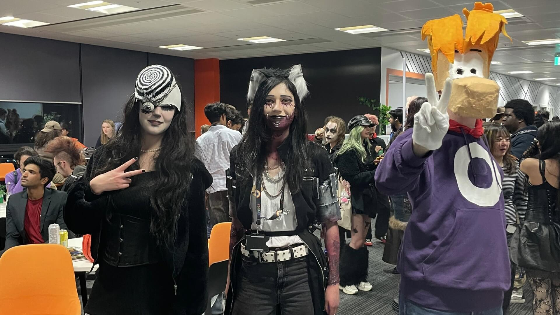Three people in Halloween costumes at an indoor event, with others in the background.