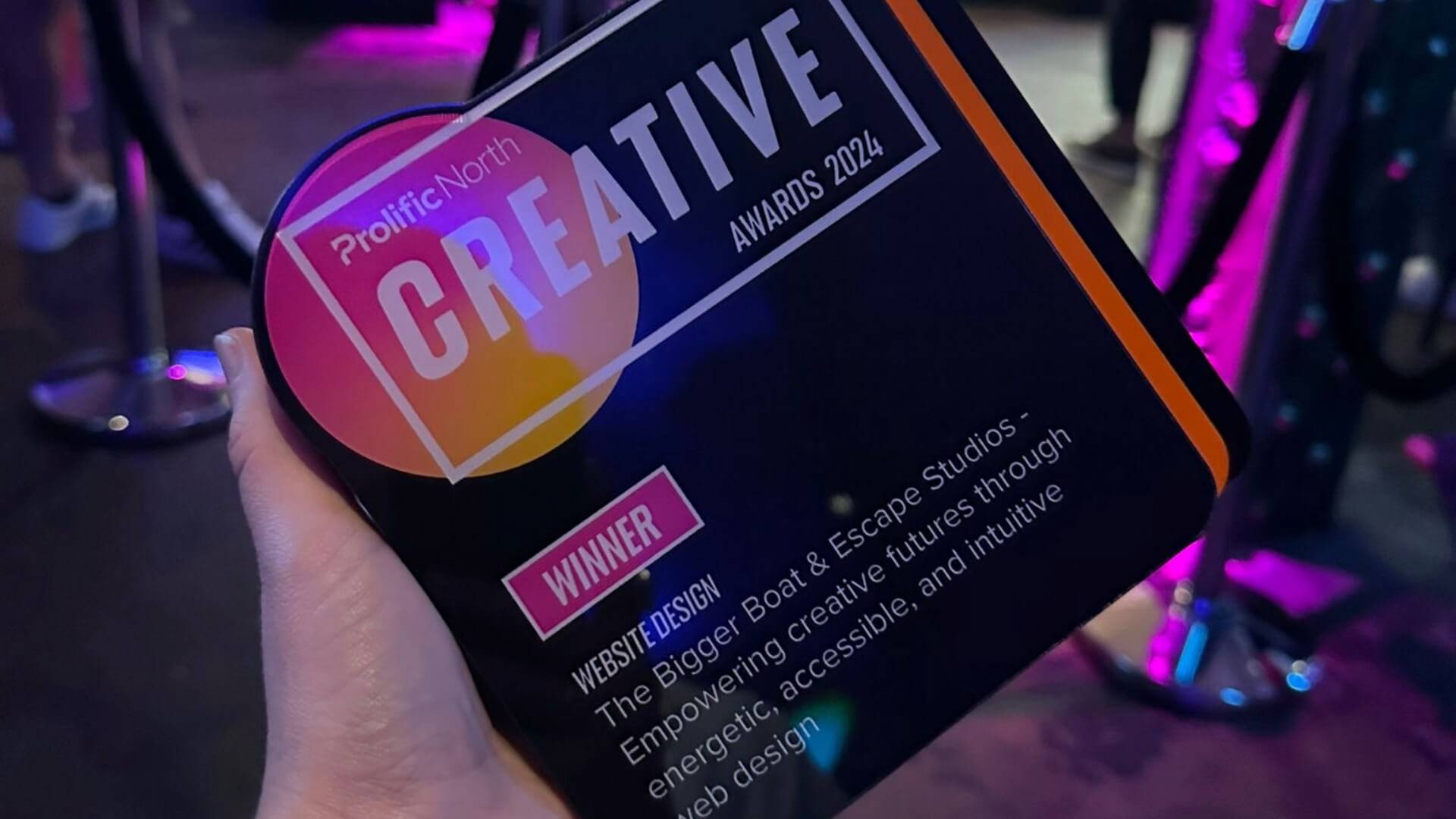 A-hand-holding-a-square-award-that-is-black-with-white-text-and-pink-and-orange-accents.-the-text-says:-'Prolific-North-Creative-awards-2024'