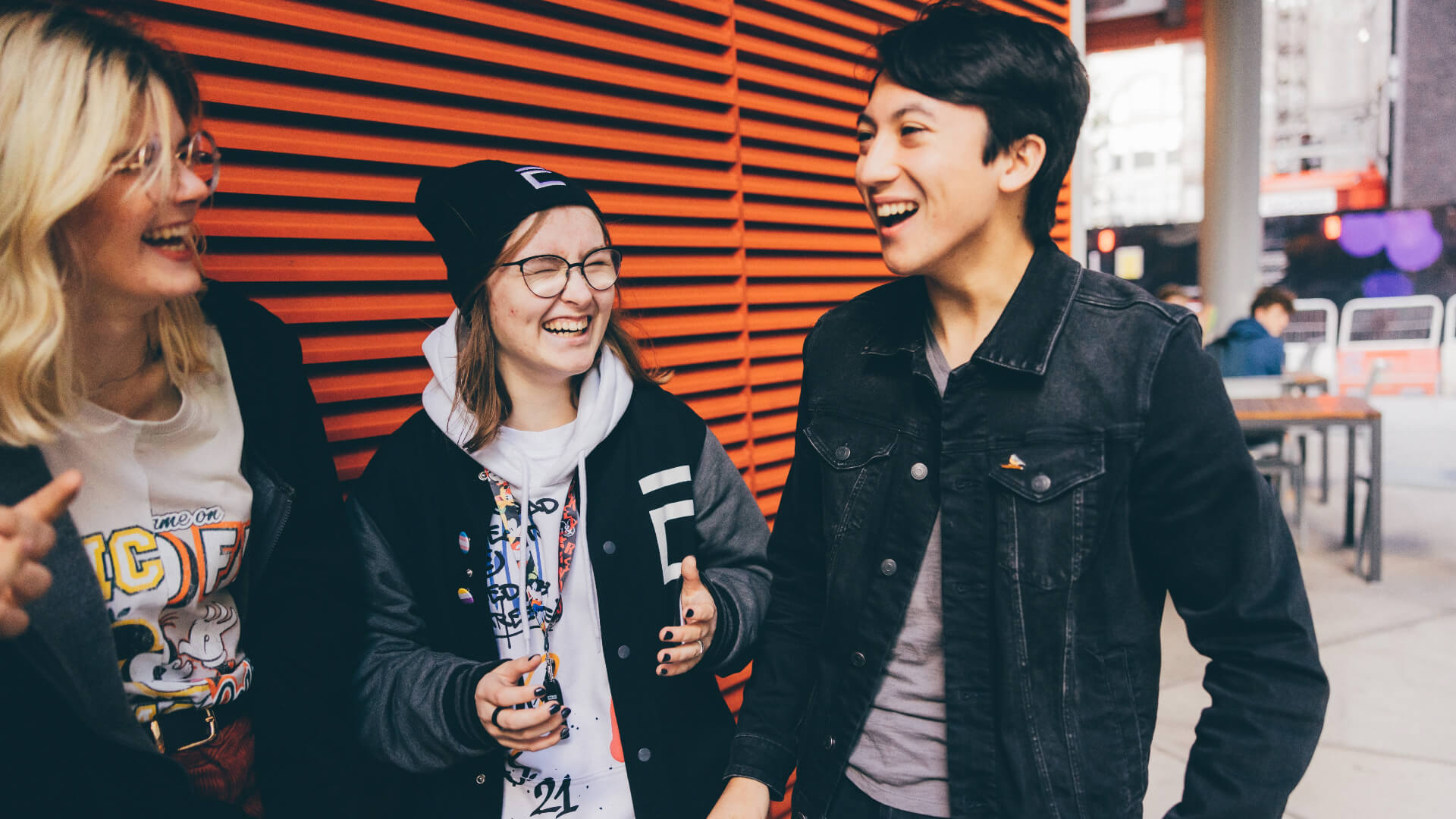 Three students laughing and talking in front of an orange wall. Kam is in the middle of the group, wearing a white hoodie.